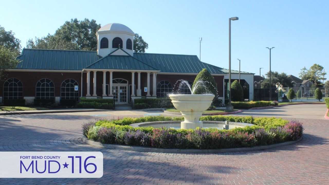 Modern community building with a central dome, fountain, and flower beds, featuring a sign for Fort Bend County MUD 116 in the foreground under a clear sky.